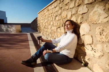 Taş bir duvarın karşısında oturmuş, güneşli bir günün tadını çıkaran, elinde kahveyle, huzur ve memnuniyeti somutlaştıran rahat bir kadın..