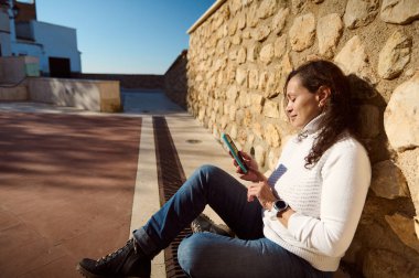 Beyaz kazaklı kadın dışarıda taş duvara karşı akıllı telefon kullanarak oturuyor. Güneşli bir gün, rahat ve nişanlı..