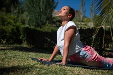 Açık hava parkında yoga yapan bir kadın, yemyeşil ve açık mavi gökyüzü ile çevrili, rahatlama ve farkındalığa odaklanmış..