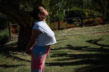 Bir kadın parkta duruyor, gülümsüyor ve güneşin sıcaklığından zevk alıyor. Rahatlamış ve mutlu görünüyor. Etrafındaki doğal güzelliğin tadını çıkarıyor..