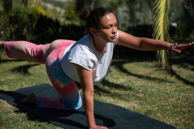 Bahçede yoga yapan bir kadın yoga minderinde denge egzersizi yapmaya odaklanmış. Güneşli bir gün, yeşil çevre sakin ve odaklanmış anı arttırıyor..