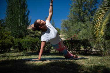 Bir kadın, güneşli bir günde yemyeşil bir bahçede paspasın üzerinde yoga pozu veriyor. Doğanın ve rahatlığın tadını çıkarıyor..