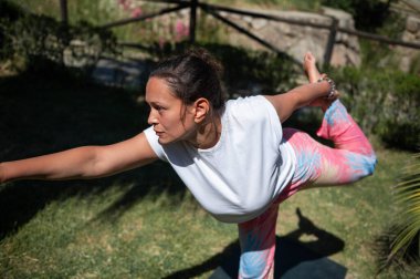 Dışarıda, sakin bir bahçe ortamında yoga yapan bir kadın Warrior III pozunu odaklanma ve konsantrasyonla veriyor. O, güneşli bir günde renkli pantolon ve beyaz bir gömlek giyiyor..