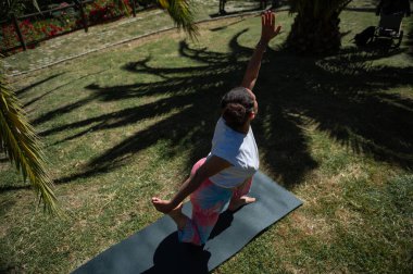 Woman doing yoga in an outdoor park setting under the sun, enjoying nature and relaxation. She engages in a stretch pose on a mat, highlighting wellness and self-care. clipart