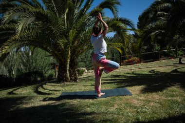 Dışarıda parkta yoga yapan bir kadın palmiye ağaçlarıyla çevrili bir şekilde ağaç pozu veriyor. Güneşli bir gün rahatlama ve farkındalık. Sağlık, meditasyon ve zindelik için uygundur..