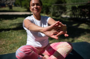 Aktif kıyafetli bir kadın, güneşli bir günde parkta yoga yaparken bacak bacak bacak üstüne atmış ve kollarını esnetiyor. Farkındalığı, rahatlığı ve sağlıklı yaşam tarzını vurgular.