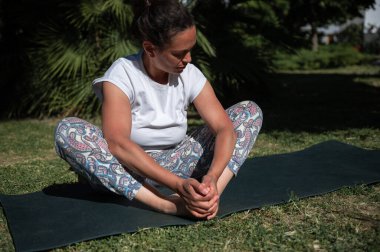 Beyaz tişörtlü ve desenli taytlı bir kadın dışarıda yoga yapıyor. Yeşillik ve güneş ışığıyla çevrili, meditasyon pozisyonunda bir mindere oturmuş..