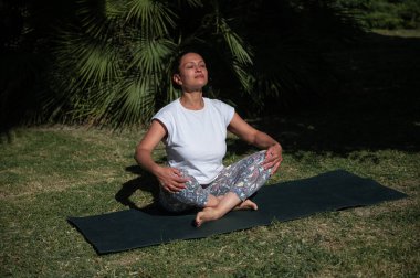 Güneşli bir açık hava parkında huzurlu bir yoga ve meditasyon anı yaşayan bir kadın sakin ve rahat bir duruşta yakalanır..