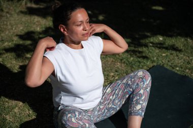 Güneşli bir bahçede yoga yapan bir kadın. Yoga minderinde oturuyor, beyaz bir bluz ve desenli taytlar giyiyor, rahatlama ve farkındalığı somutlaştırıyor..