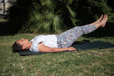 Yoga minderinde uzanmış, ayakları havada yoga pozu veren, yemyeşil bir parkta güneşli bir günün tadını çıkaran bir kadın..