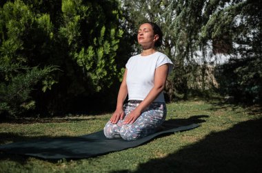 Bahçede yoga minderinde oturmuş meditasyon yapıyor ve derin nefes alıyor. Bereketli yeşillik sükuneti ve huzurlu atmosferi artırır..