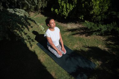 Huzurlu bir bahçedeki yoga minderinde meditasyon yapan bir kadın. Etrafı yeşilliklerle çevrili, sıcak güneş ışığı altında. İç huzuru ve sükuneti elde ediyor..