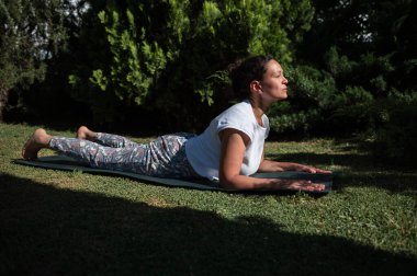 Kadın bahçede yoga yapıyor, mindere yoga pozu veriyor. Ağaçların arasından süzülen güneş ışığı huzurlu bir ortam yaratır, rahatlama ve farkındalık yaratır..