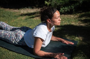 Kobrayla yoga yapan bir kadın açık havada minder üzerinde poz veriyor. Rahatlamış ve odaklanmış görünüyor, güneşli havanın ve huzurlu çevrenin tadını çıkarıyor..