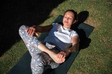 Güneşli bir günde dışarıda yoga yapan bir kadın gözleri kapalı bir mindere uzanıyor. Rahatlamayı, farkındalığı ve sağlıklı yaşam tarzını gösterir..