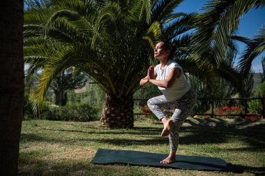 Yeşillik ve palmiye ağaçlarıyla çevrili bir parkta yoga yapan bir kadının sakin sahnesi. Odaklanma ve farkındalık ile dengeleyici bir duruş sergiliyor.