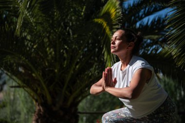Dışarıda, sakin bir tropikal ortamda yoga yapan, sabah güneşinde yıkanan bir kadın. Sakin ve odaklanmış görünüyor, yemyeşil palmiye ağaçları ve doğa ile çevrili..