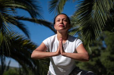 Kadın dışarıda yoga yapıyor, etrafı palmiye ağaçları ve doğa ile çevrili, sakinlik ve meditasyon anı yakalıyor. Refah, zindelik ve rahatlama temaları için idealdir.