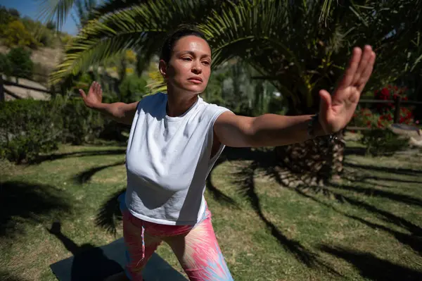 Bir kadın, yeşilliklerle çevrili güneşli bir parkta tai chi egzersizi yapıyor. Hareketlerinde konsantrasyon, denge ve farkındalık gösteriyor..