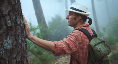 Kareli gömlekli, sırt çantalı ve şapkalı bir adam sisli bir ormanı keşfediyor. Bir ağaca dokunuyor ve doğaya odaklanmış gibi görünüyor. Macera, seyahat, keşif ve sükunet gibi kavramlar için mükemmel..