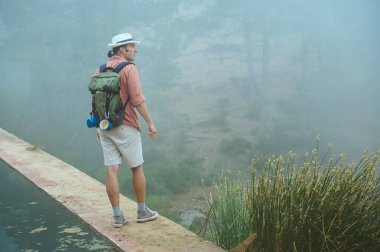 Şapka ve turuncu gömlek giyen, sırt çantası taşıyan, sisli bir orman ortamında yolda duran bir adam. Kavram: macera, doğa, keşif.