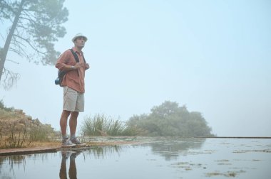 Sırt çantası takan yürüyüşçü, sisli açık havada suyun yanında duruyor ve suya yansıyor. Sahne, doğayla bir macera, yalnızlık ve bağ hissi uyandırıyor..
