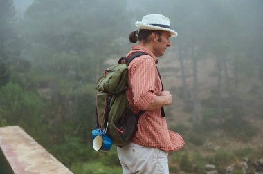 Sisli bir orman ortamında sırtında sırt çantası taşıyan, kareli gömlekli ve şapkalı bir adam. Açık hava macerası, doğa keşfi ve huzur dolu yalnızlık..