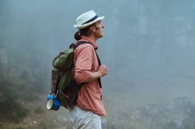 Sisli bir ormanda duran, şapka ve sırt çantası takan bir yürüyüşçü. Sahne, doğadaki macera ve keşif duygusunu yakalar..