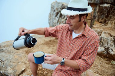 Kayalık arazide oturan bir adam şapka ve ekose gömlek giyerken termostan bardağa sıcak su döküyor. Yürüyüş, rahatlama ve doğanın tadını çıkarma kavramı..