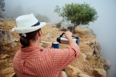 Şapkalı bir adamın, sisli bir dağ yolunda termostan sıcak su dökerken doğayı ve sükuneti kucaklayışını..