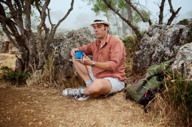 Bir adam yürüyüş sırasında yere oturmuş, etrafta kayalar ve ağaçlar varken mola veriyor. Elinde mavi bir kupa tutuyor. Doğada rahatlatıcı ve huzurlu bir an olduğunu gösteriyor..