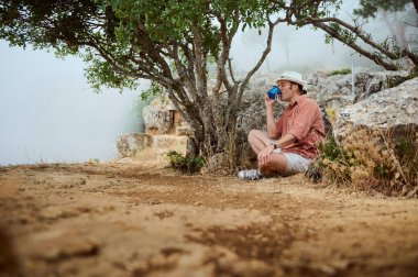 Günlük kıyafetli bir adam bir ağacın altında oturur, sakin bir açık havada kahve içer. Rahatlamayı, doğayı ve huzur dolu yalnızlığı ifade eder..