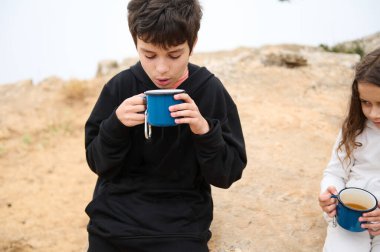 Gündelik giysiler içinde iki çocuk dışarıda oturup mavi bardaklardan sıcak içecekler içip doğanın tadını çıkarıyorlar. Görüntü rahat ve rahat bir atmosfer yakalar..