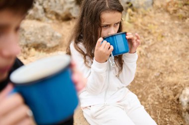Kamp yaparken mavi kupalardan içen çocukların yakın çekimi. Sahne, açık hava keşiflerinin ve çocukluk bağlarının özünü yakalıyor..