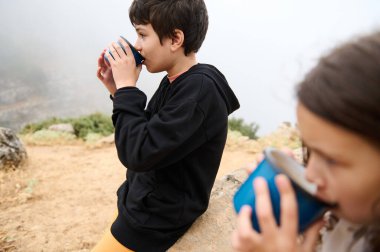 Çocuklar, açık havada sisli bir günde metal bardaklarda sıcak içeceklerin tadını çıkarıyor, açık hava maceralarının keyfini çıkarıyor ve doğayla bağ kuruyor..