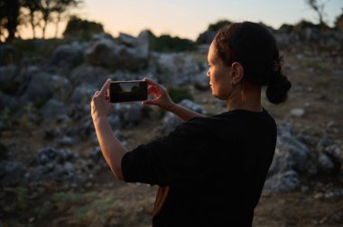 Gün batımında akıllı telefonuyla manzaralı bir anı yakalayan bir kadın. Etrafı kayalar ve doğa ile çevrili, güzel bir akşam ışığına dalmış..