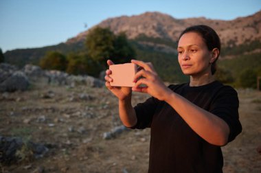 Güzel bir dağ manzarasının fotoğraflarını çekmek için akıllı telefonunu kullanan bir kadın. Güneş ışığı yüzünü hafifçe aydınlatıyor, dingin ve odaklanmış bir ifade yaratıyor..