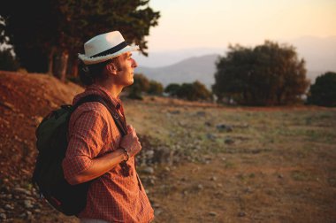 Bir adam gün batımında sırt çantasıyla yürüyüşe çıkar, şapka takar ve gömlek giyer, tepeler ve ağaçlarla manzarayı seyreder..
