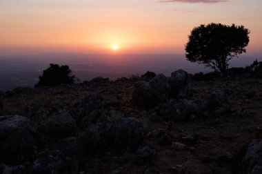Dağlık bir arazide, siluetli ağaçlar ve kayalık arazilerle baş döndürücü bir gün batımı. Gökyüzü turuncu ve mor renklerle boyanır. Huzurlu ve huzurlu bir atmosfer yaratır..