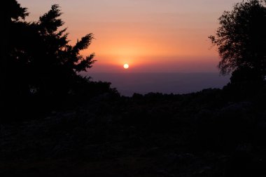 Güneşin ufukta battığı bir dağ günbatımının nefes kesici manzarası. Siluetli ağaçlar ve renkli bir gökyüzü doğa ve manzara fotoğrafçılığı için mükemmel sakin bir atmosfer yaratır..