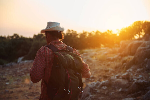 An adventurous hiker with a backpack and hat explores a rocky trail at sunset. Experience the beauty of nature and the spirit of outdoor adventure.