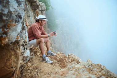 Sıradan giyinmiş bir adam, kayalık bir uçurumun kenarında oturuyor sisli bir uçuruma bakarken elinde bir bardak tutuyor. Doğanın ortasında bir anlık tefekkür.