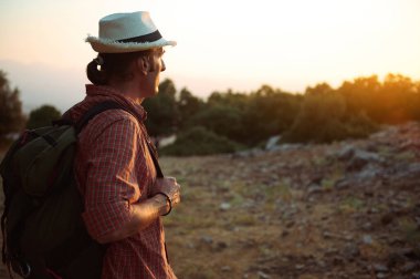 Şapkalı ve sırt çantalı bir adam güneş batarken doğa yürüyüşünden zevk alır. Macera, keşif ve sükunetin özünü yakalamak..