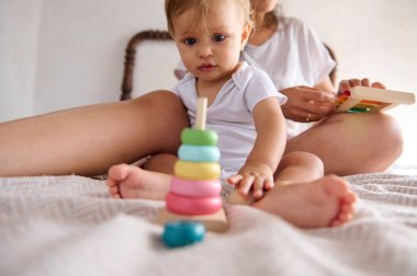 Bir bebek yatakta eğitici bir oyuncakla uğraşırken anne arka planda, oyuncu öğrenme ve gelişim sürecini ön plana çıkarıyor..