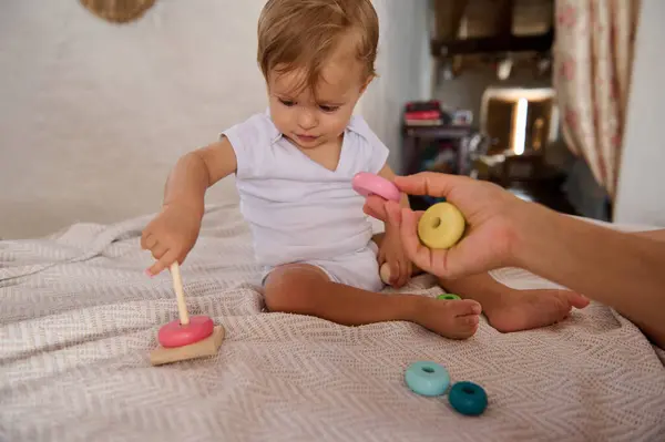 stock image Cute baby boy playing with colorful wooden stacking toy on bed, developing fine motor skills and hand-eye coordination in a cozy home environment.