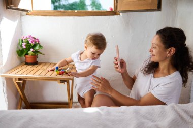 Mutlu anne, sevimli bebeğinin tahta bir masada renkli oyuncaklarla oynarken fotoğrafını çekiyor. Doğal ışık ve çiçeklerle aydınlık ve rahat bir yuva..