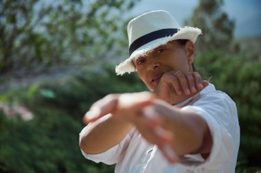 Beyaz şapkalı ve gömlekli bir kişi arka planda ağaçlarla dışarıda poz vererek sakin ve huzurlu bir atmosfer yaratıyor. Bireyler rahat ve düşünceli ve anlamlı bir duruş sergilerler..