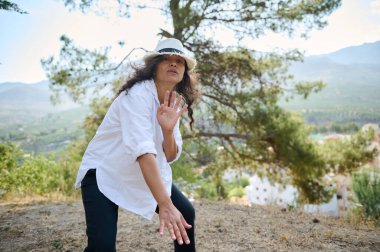 Beyaz şapkalı ve gömlekli olgun bir kadın. Açık havada kendine güvenen bir poz veriyor. Arka planda ağaçlar ve dağlar var..