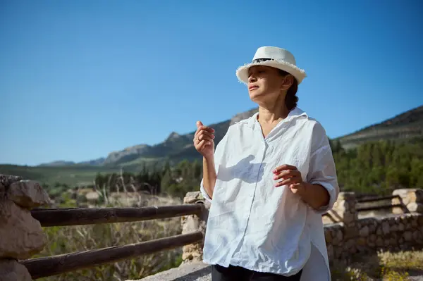 stock image Woman in white hat and blouse enjoys a sunny day in the countryside, surrounded by mountains and greenery.