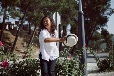 Güneşli bir günde elinde çiçekler ve ağaçlarla çevrili bir şapkayla gülümseyen neşeli bir kadın. Açık hava parkında mutluluk ve özgürlük hissi..